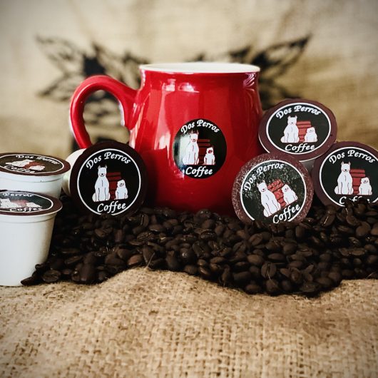 Dos Perros Coffee K cups and red mug sitting on roasted coffee beans.